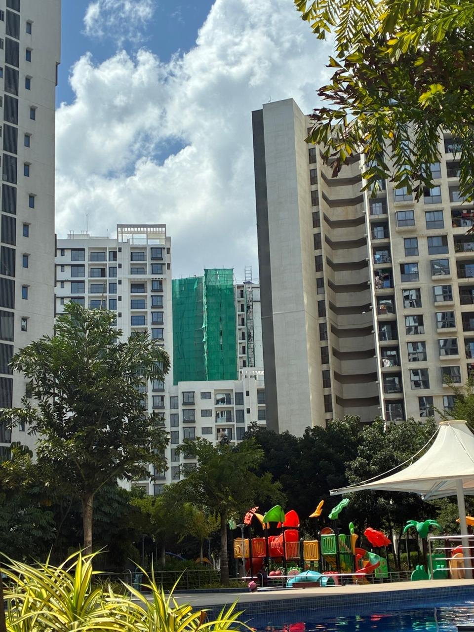 Apartment towers behind a pool and a children's play area. In the middle distance, the tower in green netting now stands visibly shorter than the towers beside it.